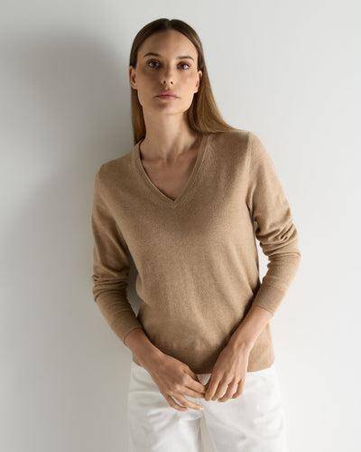 Woman in a beige V-neck knit sweater and white pants standing against a white wall.