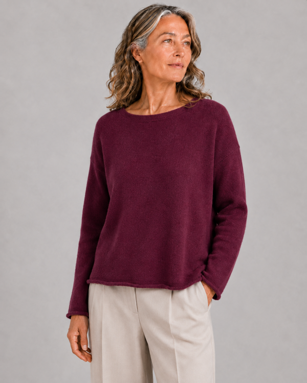 Model wearing a burgundy boat neck cashmere lambswool jumper with relaxed square shape, long sleeves and soft rolled hem finish.