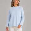 Woman in a light blue knit sweater and white pants standing against a neutral gray backdrop, looking to the side.