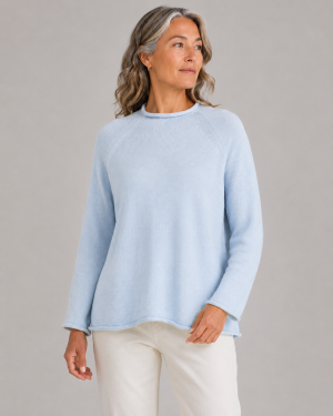 Shyla-cotton-jumper-pearl-blue-colour Woman in a light blue knit sweater and white pants standing against a neutral gray backdrop, looking to the side.