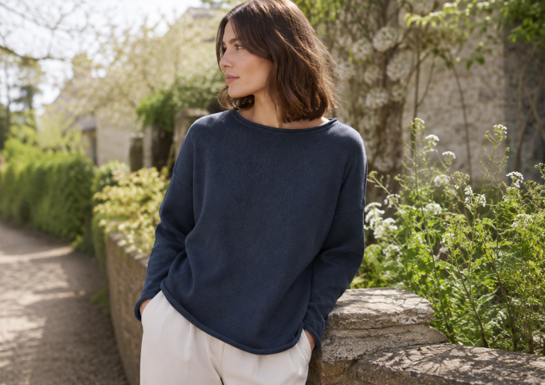 Woman leaning on a stone wall in a garden, wearing a navy sweater and light pants, looking to the left.