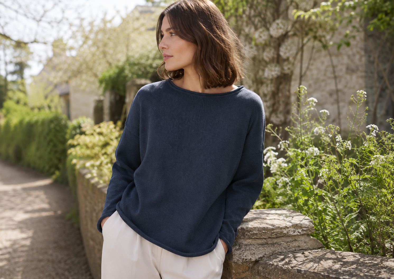 Woman leaning on a stone wall in a garden, wearing a navy sweater and light pants, looking to the left.