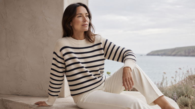 Woman wearing quiet luxury striped knitwear with cream trousers beside a soft coastal view.