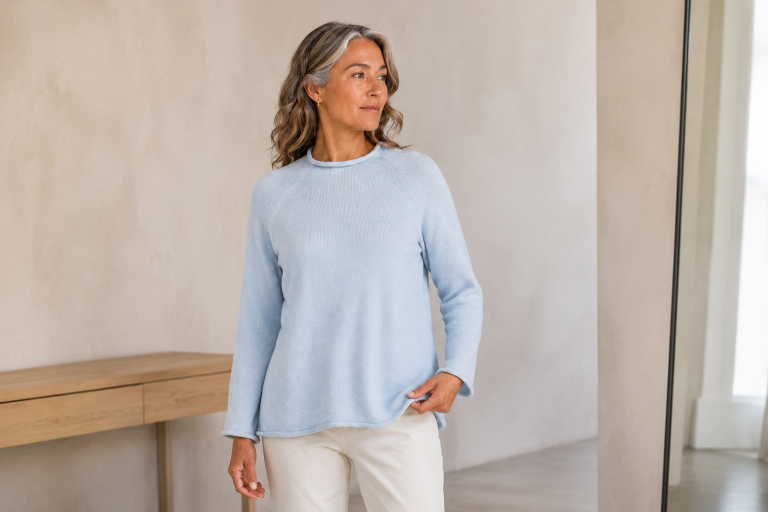 Mature woman wearing the Pearl Blue Shyla organic cotton jumper while checking its fit beside a mirror