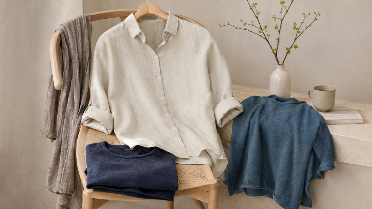 Natural fibre wardrobe weekend mood image with a linen shirt, soft knitwear layers and a scarf arranged on a wooden chair in a calm neutral interior.
