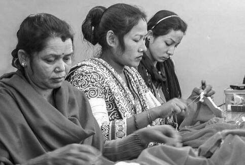 Three women sit at a table sewing fabric, focused on their handiwork in a workshop setting.