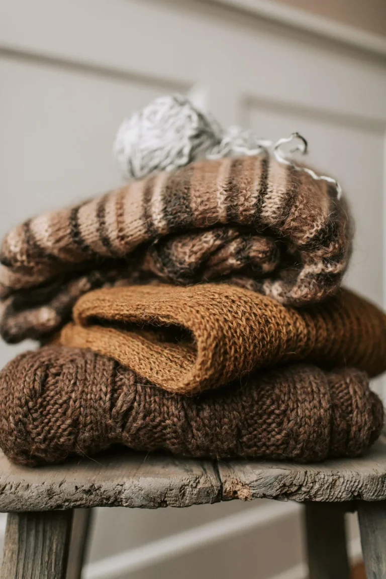 Pile of chunky knitted scarves in brown and tan tones stacked on a rustic wooden stool or table surface.