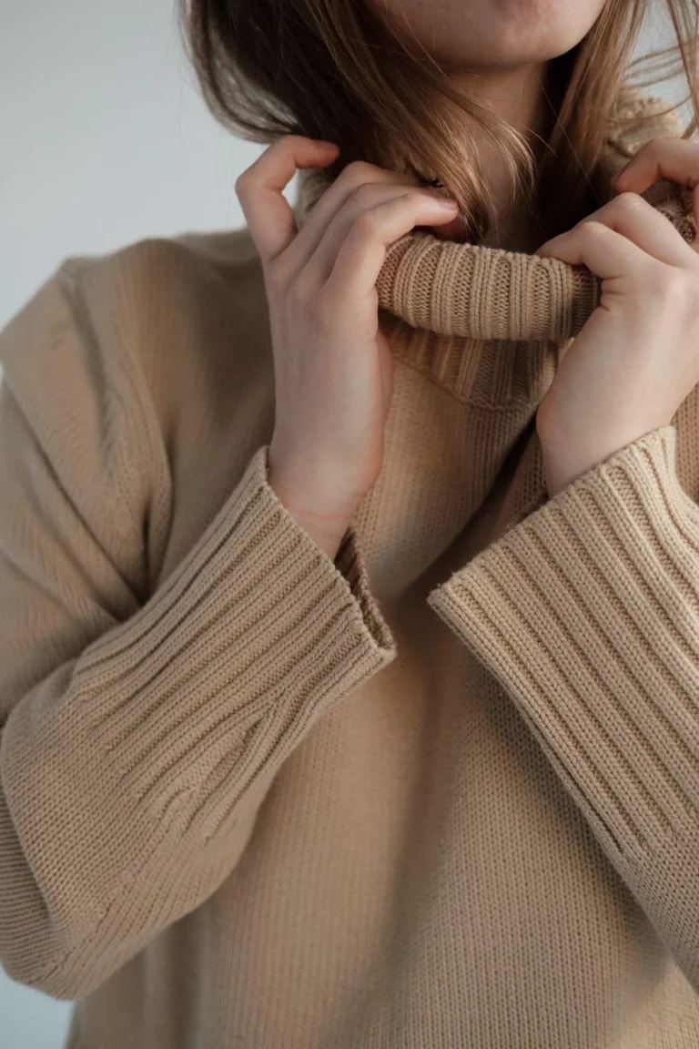 Close-up of a person in a beige knit sweater pulling the collar up at the neck.