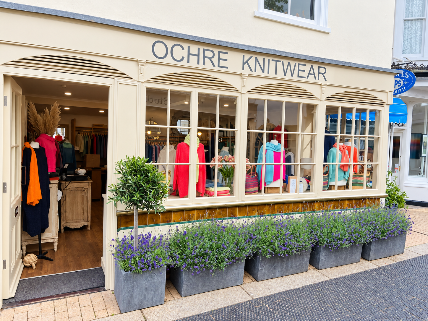 Ochre’s knitwear shop in Dartmouth with a cream shopfront, colourful window displays, an open entrance and flowering planters.