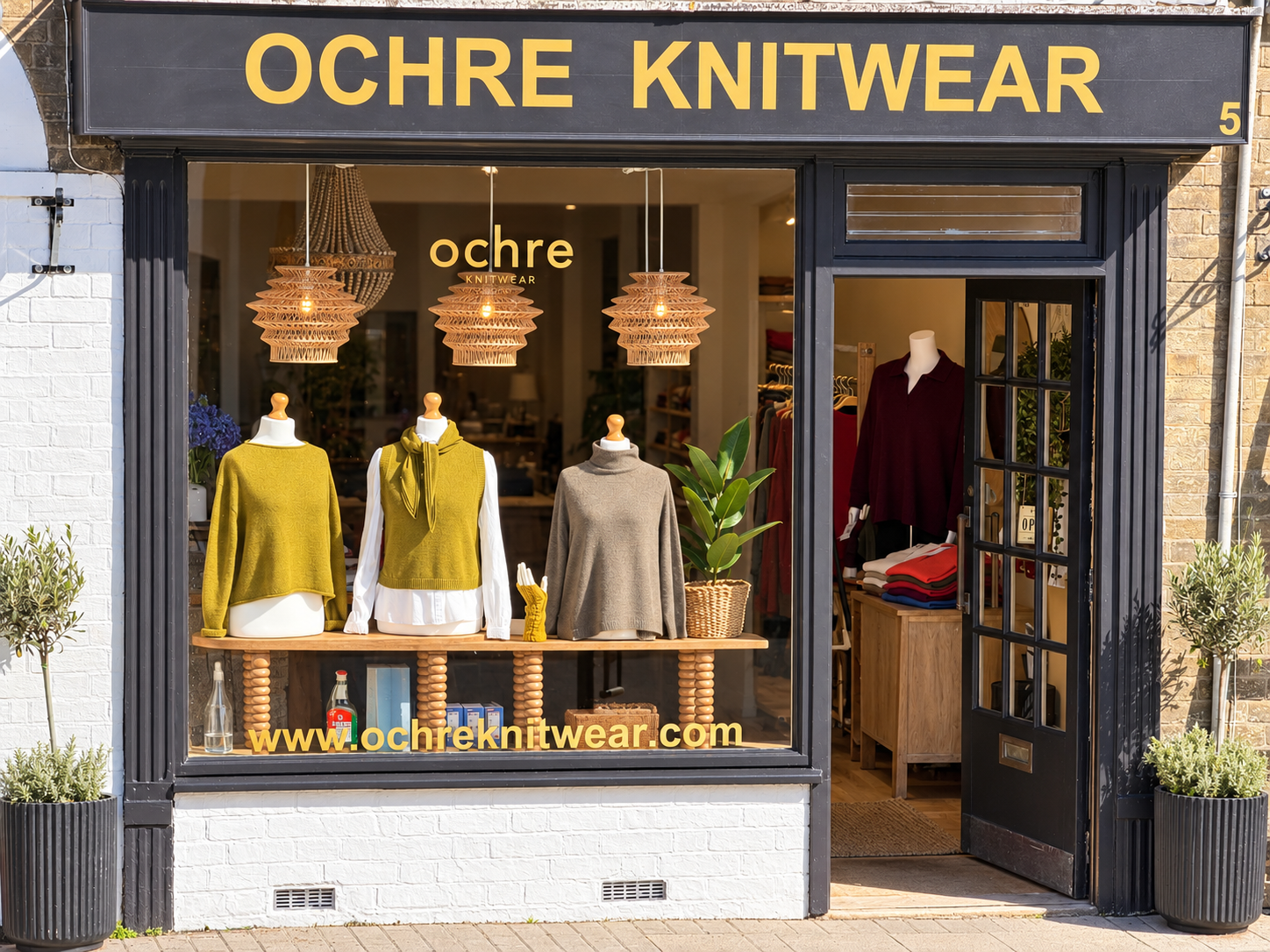Shop window display for Ochre Knitwear: three mannequins in mustard, olive, and gray sweaters under warm pendant lights, with plants inside.