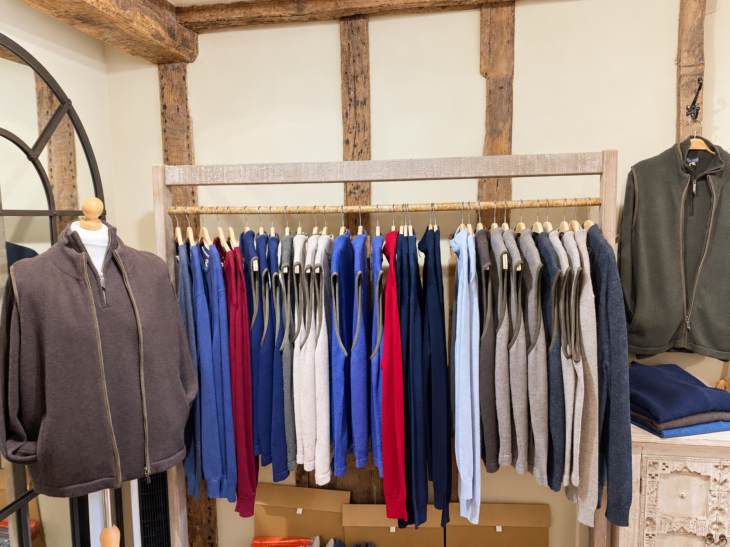 Blue, grey and cream men's knitwear on a wooden rail beneath exposed beams at Ochre’s knitwear shop in Lewes.