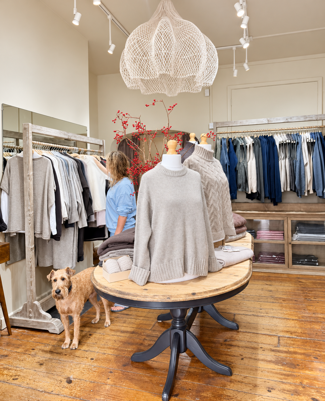 Boutique scene with beige knit sweaters on a round wooden table, clothing racks in the background, and a small dog near a shopper browsing.